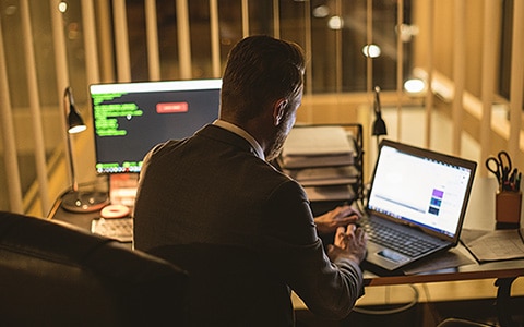 Image of a man sitting at a desk in front of his desktop computer while working on a laptop. 6 Factors Causing Cyber Insurance Rates to Increase