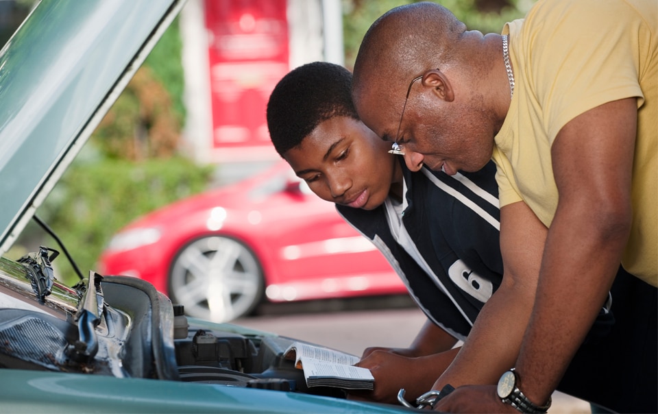 a Dad and his teenage son leaning over a car engine, reading a car maintenance manual