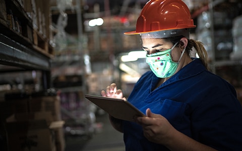 woman in mask and hard hat in a warehouse on her tablet