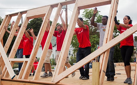 8 people 7 in red t-shirts holding up wood house frame