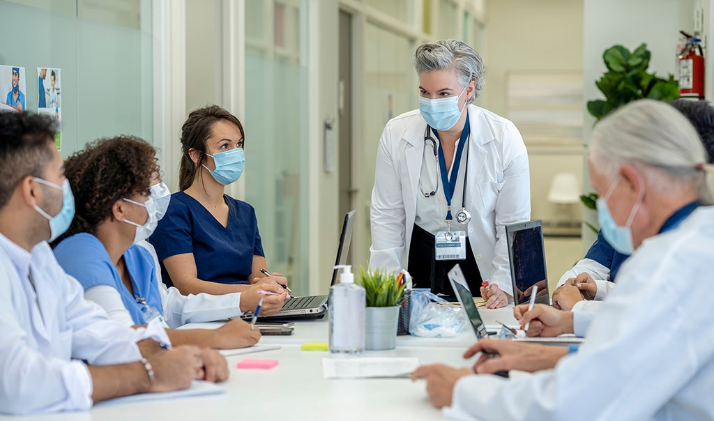 a medical professional standing in front of a table seated by other people in masks
