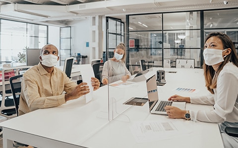 Office workers sitting at a shared workspace. They are all wearing face masks. There is a plexiglass divider in the middle of the table. How to Approach Agile Decision Making in Your Organization