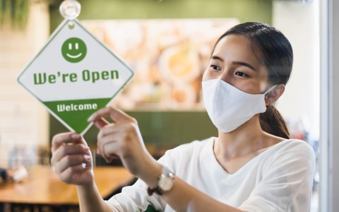 woman hanging open sign in doorway as she wears a mask