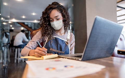 woman sitting at table at cafe in mask and apron looking at laptop and business paperwork, How to Protect Your New Business
