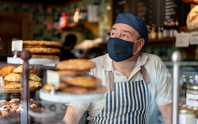 man wearing mask, hat and masks stands behinde a bakery counter, How to Scale Products and Services To Increase Profitability