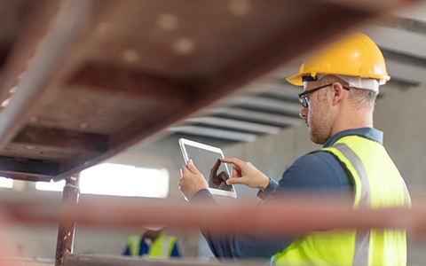 Image of a man in a hard hat working with his tablet on the job. How Interactive Safety Tools Can Help Your Business Spot and Reduce Risks