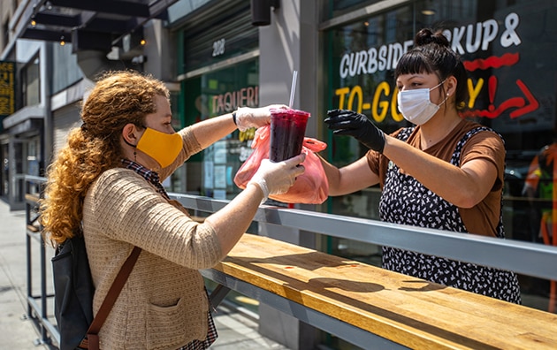 two women wearing masks and gloves. One woman is handing a restaurant to go order to another woman