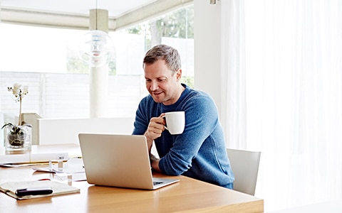 man sitting at his home table on his laptop and having a cup of coffee