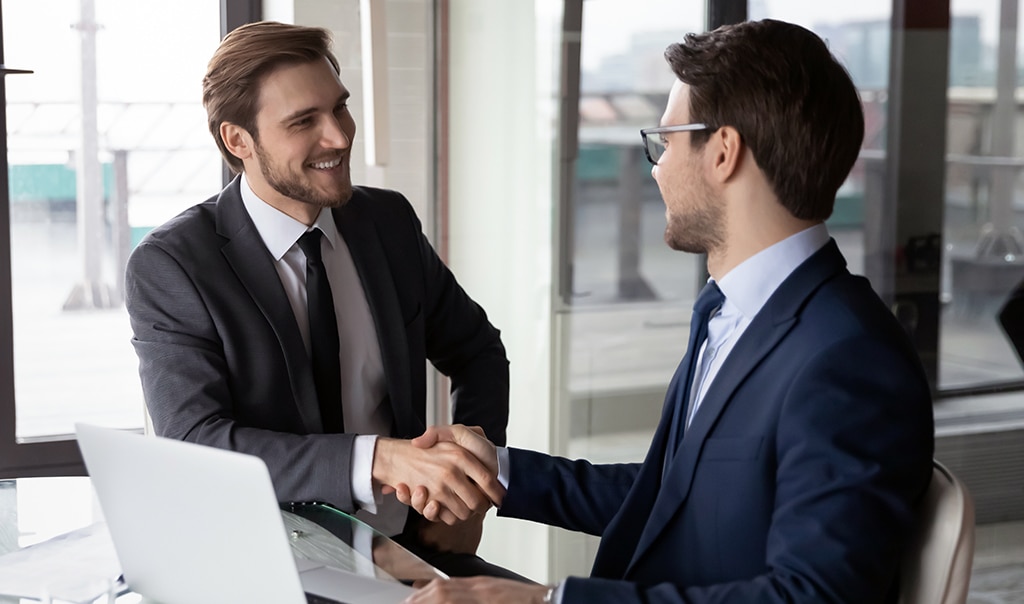 Two male professionals shaking hands, smiling, and making a deal at an office.